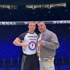 Dwóch mężczyzn w klatce MMA, jeden wskazuje na swoją koszulkę Spikes Gym Berlin
