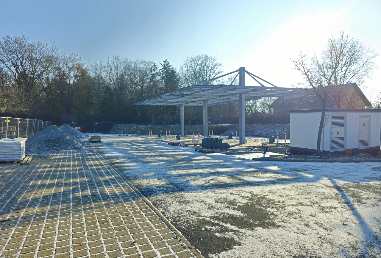 Foto reale di un parco di ricarica in costruzione: pensilina fotovoltaica su pilastri d’acciaio, cabina di trasformazione, grigliato erboso appena posato